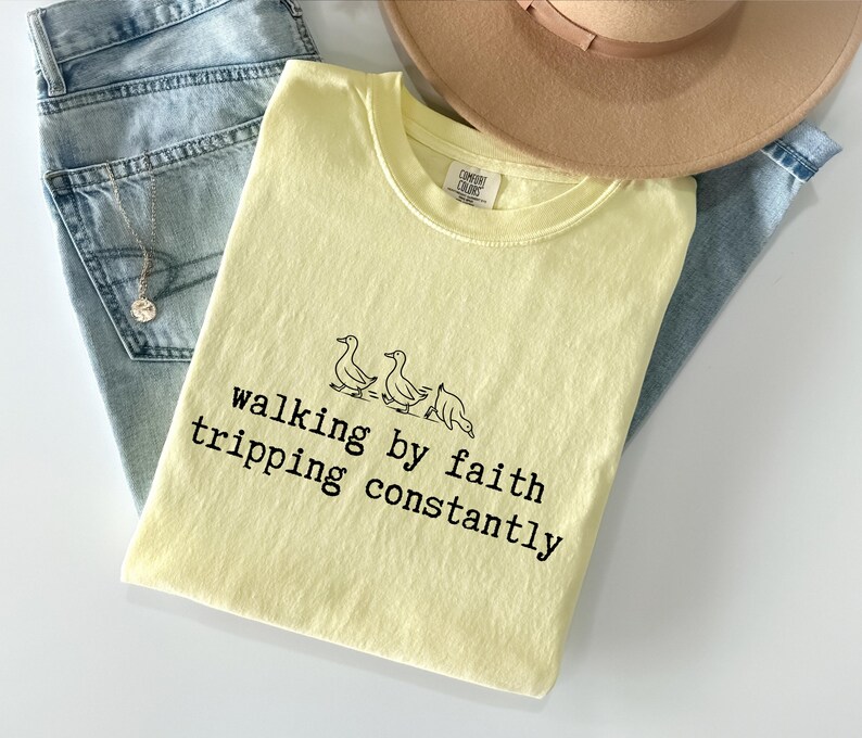 May include: A light yellow t-shirt with the text "walking by faith tripping constantly" and a graphic of three ducks. The shirt is folded on top of a pair of light blue jeans and a tan hat.
