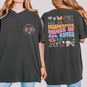 Puede incluir: Camiseta gris oscuro con el texto "NORMALIZE MINDS OF ALL KINDS" y un diseño floral en la parte delantera. La parte trasera de la camiseta presenta texto y gráficos adicionales, incluido un arcoíris y una mariposa.