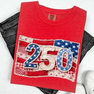 Pode incluir: Uma camiseta vermelha com um design de patchwork da bandeira americana e o número "250". O design da bandeira incorpora tecidos vermelhos, brancos e azuis. A camisa é exibida com jeans pretos e tênis brancos.