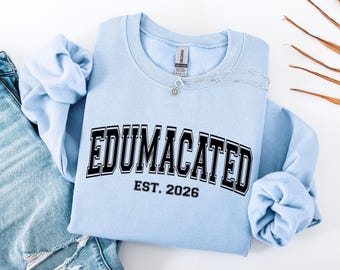 May include: A light blue crewneck sweatshirt with the word "EDUMACATED" in black block letters with a white outline. Below the word is "EST. 2026" in black. A silver necklace with a small pendant rests on the sweatshirt.