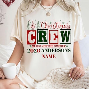 May include: A cream-colored short-sleeve t-shirt featuring a red and green graphic that reads Christmas CREW, MAKING MEMORIES TOGETHER, 2026 ANDERSONS, and NAME. The design includes small green and red Christmas tree and snowflake icons.