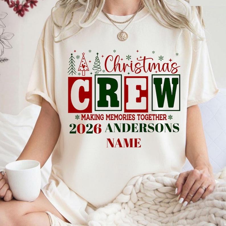 May include: A cream-colored short-sleeve t-shirt featuring a red and green graphic that reads Christmas CREW, MAKING MEMORIES TOGETHER, 2026 ANDERSONS, and NAME. The design includes small green and red Christmas tree and snowflake icons.