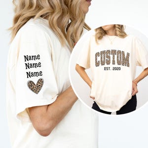 Op de afbeelding: Crèmekleurig T-shirt met het woord "CUSTOM" in een luipaardprint en de tekst "EST. 2020". De linkermouw heeft het woord "Name" drie keer herhaald, met een luipaardprint hart eronder.