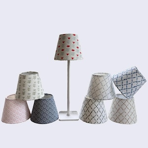 May include: A white table lamp with a red floral patterned lampshade stands among several patterned lampshades. The lampshades feature various designs, including floral, geometric, and abstract patterns in shades of pink, blue, and gray.