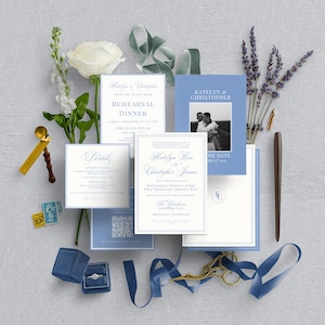 May include: An assortment of wedding stationery, including invitations, RSVP cards, and a save-the-date card, in shades of blue and white. A blue velvet ring box, a gold wax seal, and a pen are also included. The arrangement is decorated with white flowers and lavender.