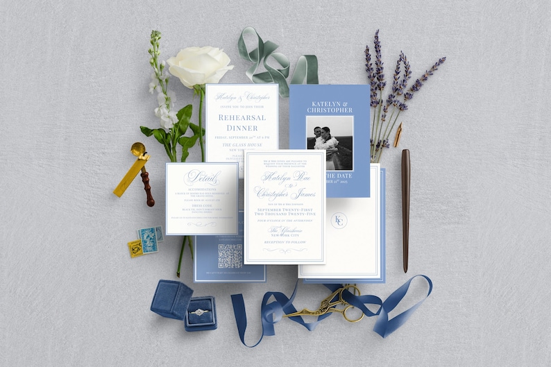 May include: An assortment of wedding stationery, including invitations, RSVP cards, and a save-the-date card, in shades of blue and white. A blue velvet ring box, a gold wax seal, and a pen are also included. The arrangement is decorated with white flowers and lavender.