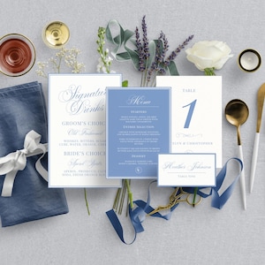 May include: A flat lay of wedding stationery and table settings in shades of blue, white, and gold. Includes menu cards, place cards, a napkin, and silverware. The table number card displays the number 1. The overall theme is elegant and classic.