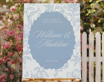 May include: A wedding welcome sign on a wooden easel. The sign features a light blue oval with the names "William & Madeline" in elegant script. The background is white with a floral pattern, and the date 06.16.2026 is displayed.