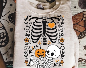 May include: Beige sweatshirt featuring a black skeleton design with a heart, flowers, and a pumpkin. The skeleton is holding a pumpkin. The text "PNG - SVG - EPS" is at the bottom. A Halloween-themed garment.