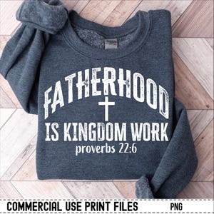 May include: A dark gray sweatshirt with the words "FATHERHOOD IS KINGDOM WORK" in white, along with a cross and the Bible verse Proverbs 22:6. The sweatshirt is folded on a light brown wooden surface.