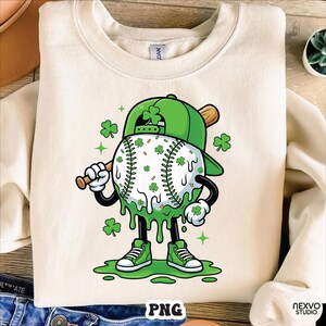 K&ouml;nnte beinhalten: Cremefarbenes Sweatshirt mit einem Baseball-Charakter zum St. Patrick's Day. Der Baseball ist wei&szlig; mit gr&uuml;nen Kleebl&auml;ttern und gr&uuml;nen Tropfenakzenten. Die Figur tr&auml;gt eine gr&uuml;ne Baseballkappe und gr&uuml;ne Turnschuhe und h&auml;lt einen Holzschl&auml;ger.