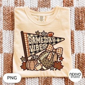May include: A light yellow t-shirt featuring a graphic of a pennant flag with the text GAME DAY VIBES, a football, a pumpkin, a large bow, and autumn leaves. The design incorporates plaid, leopard print, and star patterns in brown and black tones.
