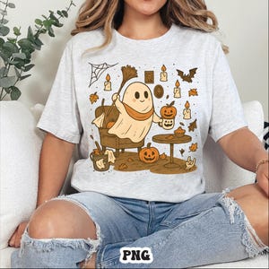 May include: A light gray t-shirt featuring a Halloween-themed graphic. The design includes a cartoon ghost holding a broom, pumpkins, candles, and autumn leaves. The ghost is wearing an orange scarf. The shirt has a crew neck.