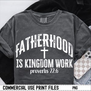 May include: Dark gray long-sleeve t-shirt with the text "FATHERHOOD IS KINGDOM WORK" and a cross graphic. The text "proverbs 22:6" is below the text. The shirt is made of a soft material.