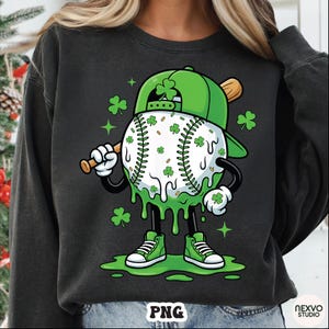 K&ouml;nnte beinhalten: Schwarzes Sweatshirt mit einer Cartoon-Baseballfigur, die eine gr&uuml;ne Kappe tr&auml;gt und einen Schl&auml;ger h&auml;lt. Der Baseball ist mit Kleebl&auml;ttern verziert, und die Figur hat gr&uuml;ne Turnschuhe und tropfenden gr&uuml;nen Schleim.