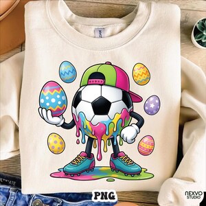May include: Cream-colored sweatshirt featuring a cartoon soccer ball character wearing a baseball cap and soccer cleats. The soccer ball is decorated with colorful dripping paint and surrounded by Easter eggs. The word "PNG" is visible.