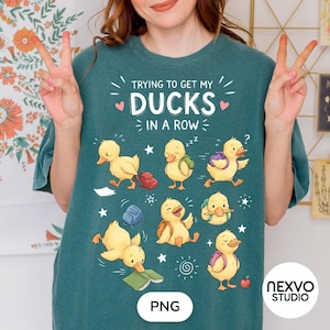 Può includere: Una t-shirt verde acqua scuro con una grafica di otto anatroccoli gialli con materiale scolastico e il testo "TRYING TO GET MY DUCKS IN A ROW" in lettere bianche, circondato da piccoli cuori rosa, stelle e spirali.