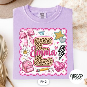 May include: A light purple t-shirt featuring a graphic print of a large leopard print letter E with the name Emma in pink script. The design includes a pink gingham bow, a daisy, a pink backpack, a pencil, a star, a heart, and a lightning bolt.