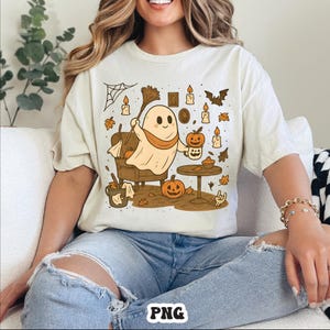 May include: Cream-colored t-shirt featuring a cartoon ghost with a scarf, holding a broom and a pumpkin mug. The design includes pumpkins, candles, and autumn leaves, creating a Halloween theme. The shirt is a relaxed fit.