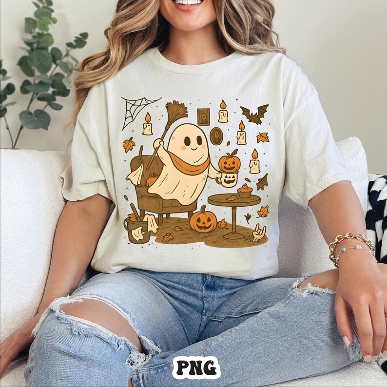 May include: Cream-colored t-shirt featuring a cartoon ghost with a scarf, holding a broom and a pumpkin mug. The design includes pumpkins, candles, and autumn leaves, creating a Halloween theme. The shirt is a relaxed fit.