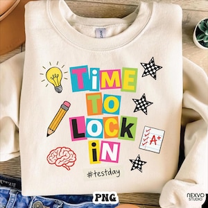 Puede incluir: Sudadera color crema con la frase "TIME TO LOCK IN" en letras de bloque coloridas. El diseño incluye una bombilla, un lápiz, un cerebro, estrellas y un examen con una calificación A+. También se incluye el hashtag #testday.