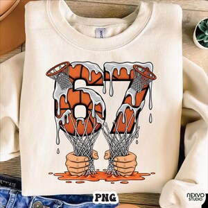 May include: Cream-colored sweatshirt featuring a basketball-themed graphic. The number "67" is designed with basketballs and dripping white paint. Two hands are holding basketball nets, with orange paint at the bottom. The word "PNG" is at the bottom.