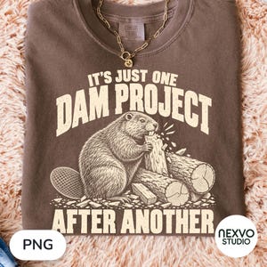 Könnte beinhalten: Ein braunes T-Shirt mit einer Grafik eines Bibers, der an einem Holzstamm nagt, und dem cremefarbenen Schriftzug IT'S JUST ONE DAM PROJECT AFTER ANOTHER. Das T-Shirt hat einen Rundhalsausschnitt und eine goldfarbene Kette liegt auf dem Stoff.
