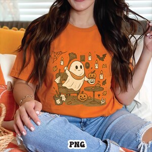 May include: Orange t-shirt featuring a cartoon ghost with a broom, holding a pumpkin-shaped mug. The design includes pumpkins, candles, bats, and a spiderweb, perfect for Halloween. The shirt is a comfortable fit.