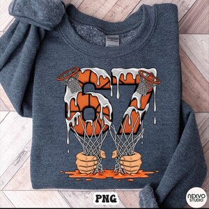 May include: A dark gray sweatshirt featuring a basketball-themed graphic. The number "67" is designed with basketballs and nets, with a dripping white and orange effect. The word "PNG" is at the bottom.