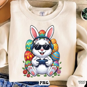 Könnte beinhalten: Cremefarbenes Sweatshirt mit einem Cartoon-Osterhasen, der eine Sonnenbrille und Kopfhörer trägt und einen Gamecontroller hält. Der Hase ist von bunten Ostereiern und Blumen umgeben. Das Bild hat unten den Text "PNG".