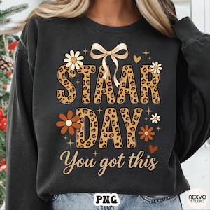 May include: Dark grey sweatshirt featuring the text "STAAR DAY" in leopard print lettering, decorated with floral and heart graphics. The phrase "You got this" is written in a cursive font. A bow graphic is at the top.