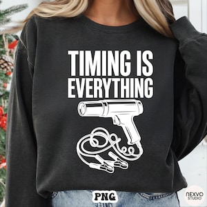 Puede incluir: Sudadera gris oscuro con el texto blanco "TIMING IS EVERYTHING". Debajo, un gráfico blanco de una pistola de calor con cables y pinzas. La sudadera es de estilo casual.