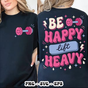 Op de afbeelding: Zwart T-shirt met een roze halter-afbeelding en de woorden "Be Happy Lift Heavy" in roze en blauw. Het ontwerp bevat sterren, bliksemschichten en waterdruppels. Het shirt is gemaakt van zacht materiaal.