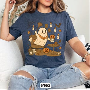 May include: Navy blue t-shirt featuring a cartoon ghost with a broom, pumpkins, candles, and autumn leaves. The ghost is holding a cup and sitting in a chair. The shirt has a Halloween theme.