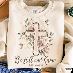 Könnte beinhalten: Cremefarbenes Sweatshirt mit einem Kreuzdesign mit Leopardenmuster-Umrandung, umgeben von floralen und Blättern. Der Text "Be still and know" und "Psalm 46:10" sind darunter gedruckt. Zwei Tauben sind ebenfalls abgebildet.
