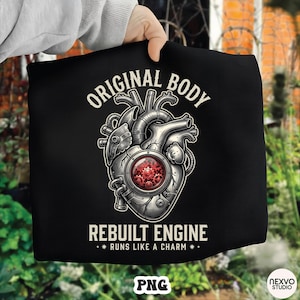 Pode incluir: Camiseta preta com uma ilustração detalhada de um coração mecânico. O design inclui o texto "ORIGINAL BODY" acima do coração e "REBUILT ENGINE" abaixo, com "RUNS LIKE A CHARM" no meio.