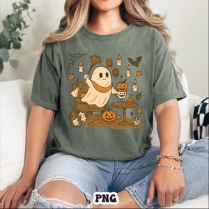 May include: Sage green t-shirt featuring a cartoon ghost with a pumpkin mug, surrounded by Halloween-themed illustrations. The design includes pumpkins, candles, and autumn leaves, creating a festive and whimsical vibe. The shirt is a comfortable, casual style.