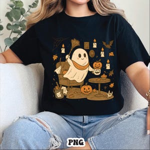 May include: Black t-shirt featuring a cartoon ghost holding a broom and a pumpkin-shaped mug. The design includes pumpkins, candles, and autumn leaves, creating a Halloween theme. The shirt has a relaxed fit.