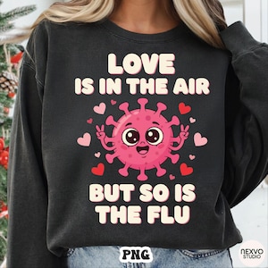 May include: Dark gray sweatshirt with the text "LOVE IS IN THE AIR BUT SO IS THE FLU". The design features a pink cartoon virus with a smiling face, peace signs, and hearts.
