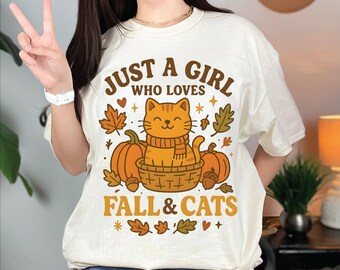 May include: Cream-colored t-shirt with the text "JUST A GIRL WHO LOVES FALL & CATS." The graphic features a smiling orange cat wearing a scarf, sitting in a basket with pumpkins and fall leaves. The shirt has a relaxed fit.