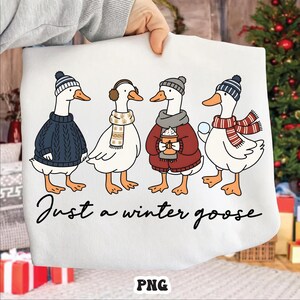 May include: White sweatshirt featuring four cartoon geese dressed for winter. Each goose wears a hat, scarf, or earmuffs. One goose holds a snowball, and another holds a mug. The text "Just a winter goose" is written below the geese.