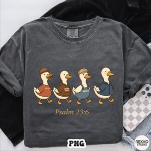May include: Dark gray long-sleeve shirt featuring a cartoon illustration of four ducks wearing different outfits. The ducks are walking in a row, with the text "Psalm 23:6" below them. The shirt has a vintage, washed look.
