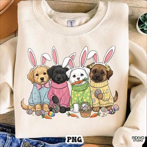 May include: Cream-colored sweatshirt featuring a graphic of four puppies dressed as Easter bunnies. Each puppy wears bunny ears and a sweater, with Easter eggs and carrots scattered around them. The word "PNG" is at the bottom.