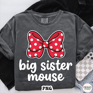 May include: Dark gray long-sleeve shirt featuring a large red bow with white polka dots. The words "big sister mouse" are printed in white below the bow. A phone with a checkered case is visible in the background.