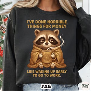 Puede incluir: Sudadera gris oscuro con el texto "I'VE DONE HORRIBLE THINGS FOR MONEY LIKE WAKING UP EARLY TO GO TO WORK." Un mapache de dibujos animados sostiene una taza con vapor saliendo. El mapache está envuelto en una manta.