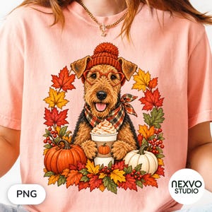 Puede incluir: Una camiseta color melocotón con el gráfico de un Airedale Terrier que lleva un gorro de punto rojo, gafas en forma de corazón y una bufanda de cuadros. El perro sostiene un café con nata, rodeado de hojas de otoño, bayas y calabazas.