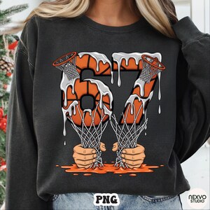 May include: Dark gray sweatshirt featuring a basketball-themed graphic. The design includes the number "67" formed by basketballs with dripping white paint, basketball nets, and hands holding the nets. The overall design has an orange and white color scheme.