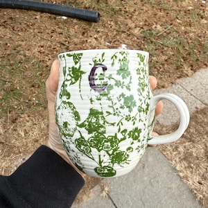 May include: White ceramic mug with a green floral pattern and a purple letter "C". The mug has a curved handle and is held by a person. The mug is cylindrical and appears to be handmade.