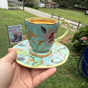 Puede incluir: Un juego de taza y platillo de cerámica turquesa con diseños florales y un borde amarillo. La taza tiene un asa única y está llena de un líquido dorado. Un pequeño letrero que dice "Welcome friends" está en el fondo.