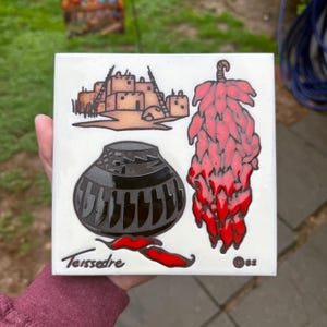 May include: A white ceramic tile with a red chili pepper, a black pot with a geometric design, and a brown adobe building with a ladder. The tile is signed "Teissedre" and "82".